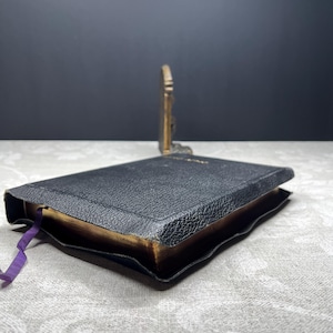 Puede incluir: Un libro peque&ntilde;o, negro, con una cubierta texturizada y p&aacute;ginas doradas. Una cinta morada como marcador de libros sobresale del libro. Un objeto decorativo de color dorado est&aacute; en el fondo. El libro est&aacute; ligeramente abierto.