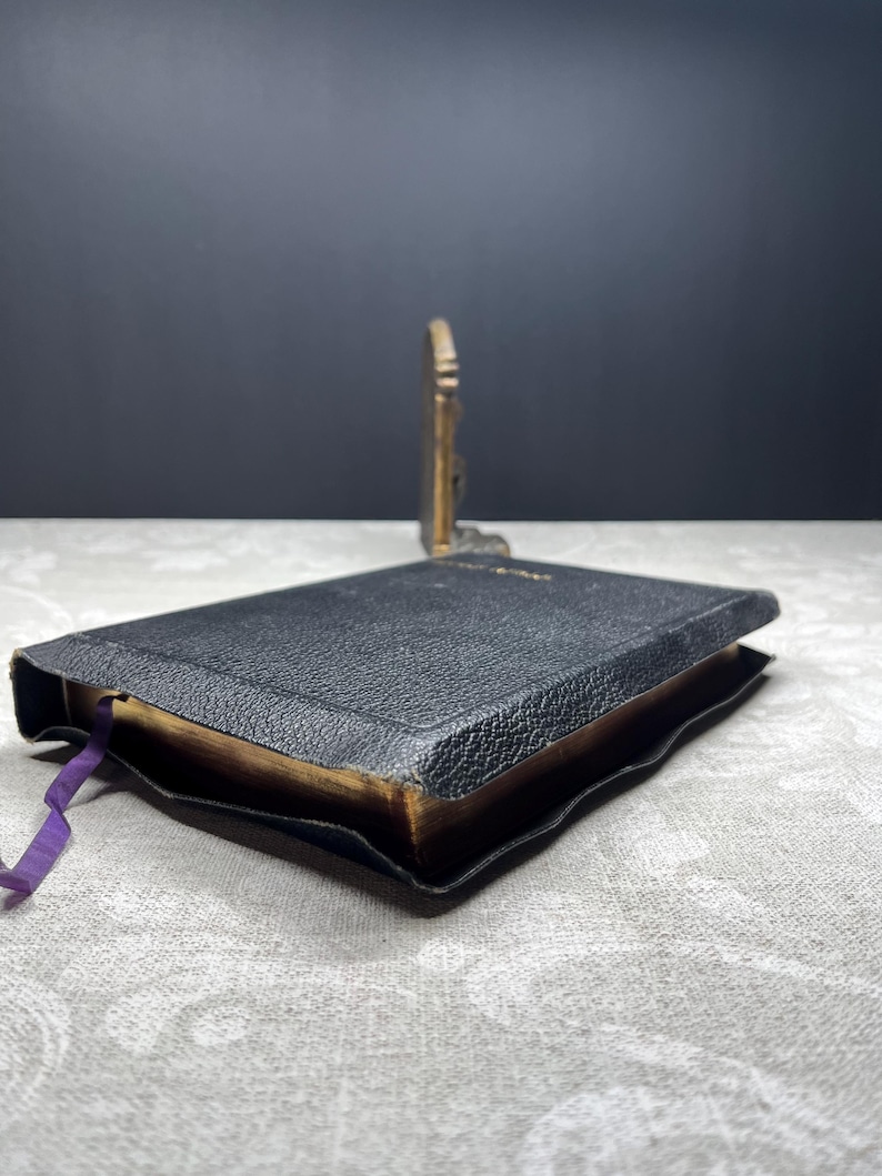 Puede incluir: Un libro peque&ntilde;o, negro, con una cubierta texturizada y p&aacute;ginas doradas. Una cinta morada como marcador de libros sobresale del libro. Un objeto decorativo de color dorado est&aacute; en el fondo. El libro est&aacute; ligeramente abierto.