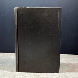 May include: A vintage hardcover book with a dark, textured cover. The book's spine is visible, showing signs of wear. The edges of the pages are slightly aged, suggesting the book's age. The book is standing upright, showcasing its rectangular shape.