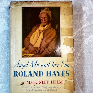 Op de afbeelding: Vintage boekomslag met een portret van een oudere persoon met een gele sjaal. De titel "Angel Mo' and her Son Roland Hayes" staat in het blauw, met de naam van de auteur, Mackinley Helm.