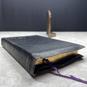 Puede incluir: Un libro encuadernado en cuero negro con bordes dorados y una cinta morada como marcador. El libro est&aacute; ligeramente desgastado, con texto visible en el lomo. Un peque&ntilde;o objeto decorativo dorado se encuentra en el fondo.