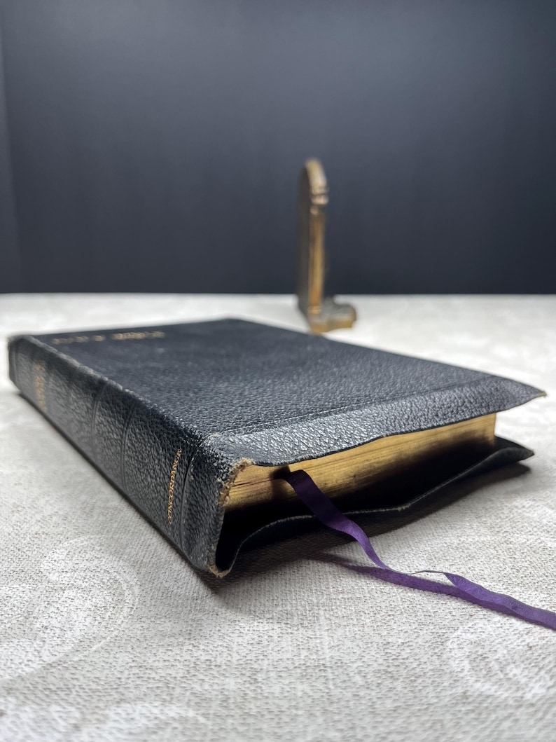 Puede incluir: Un libro encuadernado en cuero negro con bordes dorados y una cinta morada como marcador. El libro est&aacute; ligeramente desgastado, con texto visible en el lomo. Un peque&ntilde;o objeto decorativo dorado se encuentra en el fondo.