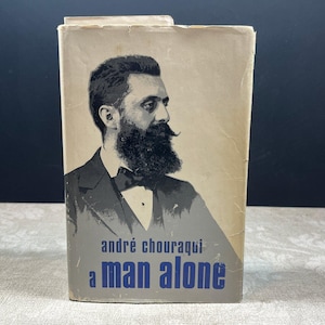 May include: A hardcover book with a cream and gray dust jacket featuring a black and white portrait of a bearded man in a suit. The text on the cover reads "andré chouraqui" in small black letters and "a man alone" in large blue letters.