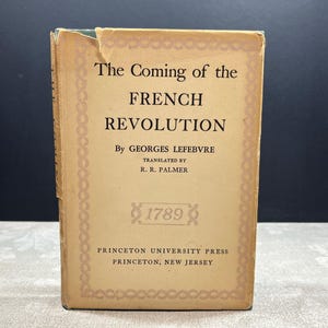 May include: A vintage book titled "The Coming of the French Revolution" by Georges Lefebvre, translated by R.R. Palmer. The cover is tan with black text and decorative borders. The year 1789 is also printed on the cover. Published by Princeton University Press.