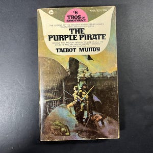 Puede incluir: Un libro de bolsillo titulado The Purple Pirate de Talbot Mundy, parte de la serie Tros of Samothrace. La portada presenta una escena ilustrada oscura con figuras en un barco y una sección superior de color crema que contiene texto.