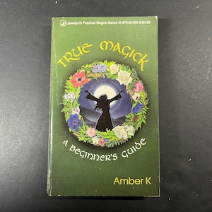 May include: A paperback book titled True Magick: A Beginner's Guide by Amber K. The dark green cover features a central illustration of a silhouette figure with arms raised against a full moon, surrounded by a colorful floral wreath of roses.