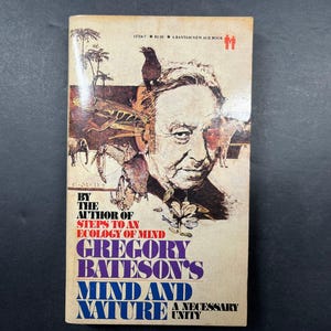 Op de afbeelding: Een paperbackboek getiteld Mind and Nature: A Necessary Unity van Gregory Bateson. De kaft bevat een sepia-kleurige illustratie van een mansgezicht omringd door diverse dieren en planten, met tekst in zwarte, rode en blauwe inkt.