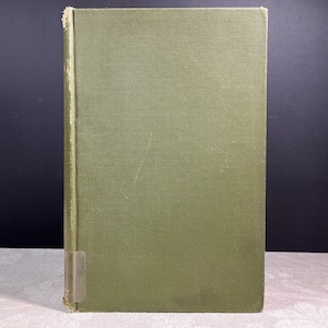 May include: A vintage hardcover book with a textured olive green cover. The spine shows wear and a small label. The book is standing upright against a dark background, suggesting a focus on its presentation.