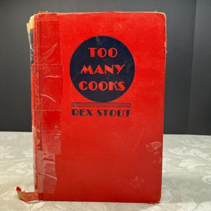 May include: A vintage hardcover book titled "Too Many Cooks" by Rex Stout. The book has a bright red cover with a navy blue circle containing the title in white text. The book is wrapped in clear plastic.