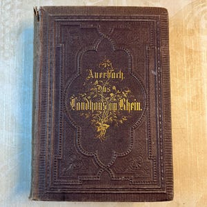 May include: A brown leather-bound book with gold lettering on the cover. The title is "Auerbach, Das Landhaus am Rhein".