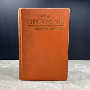 以下が含まれることがあります： J. Johnston Abrahamによる「The Surgeon's Log」というタイトルの古い本。オレンジ色の表紙に金色の文字が書かれています。本は直立しています。
