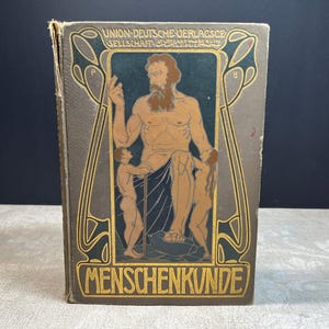 May include: A hardcover book titled MENSCHENKUNDE with a grey cover featuring a gold and black illustration of a central bearded figure flanked by two smaller figures. The text UNION DEUTSCHE VERLAGSGESELLSCHAFT STUTTGART BERLIN LEIPZIG is printed at the top.