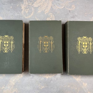 Pode incluir: Três livros de capa dura verde com detalhes dourados. Cada livro apresenta um brasão dourado com uma cruz e uma coroa na capa.