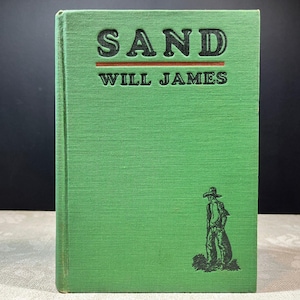 May include: A green hardcover book titled SAND by Will James. The front cover features black embossed text, a horizontal red line, and a black line drawing of a person wearing a wide-brimmed hat standing on a patch of ground.