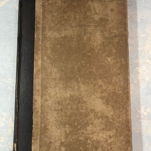 May include: A vintage book with a worn, tan cover and a black spine. The cover shows signs of age with discoloration and wear. The book's spine is black with a textured appearance. The overall aesthetic suggests a well-loved, antique item.