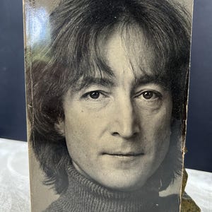 Puede incluir: Una portada de libro vintage en blanco y negro con un retrato de John Lennon. La portada tiene el título "Strawberry Fields Forever: John Lennon Remembered" en una fuente serif. La imagen es un primer plano del rostro de Lennon.
