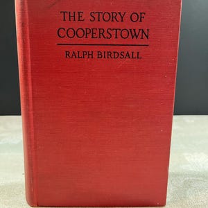 May include: A red hardcover book titled "The Story of Cooperstown" by Ralph Birdsall. The title and author's name are printed in black on the front cover. The book is standing upright.
