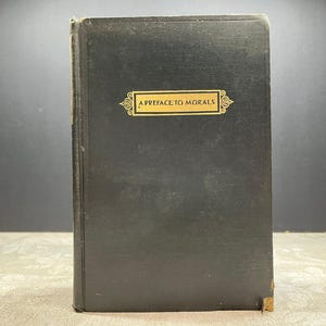 Pode incluir: Um livro antigo de capa dura com capa preta. O título "A Preface to Morals" é exibido em letras douradas em um rótulo retangular. O livro mostra sinais de desgaste, com bordas ligeiramente desgastadas.