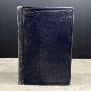 Op de afbeelding: Een vintage, donkerblauw boek met gouden letters op de rug. De kaft heeft een textuur en decoratieve hoekontwerpen. Het boek staat rechtop tegen een donkere achtergrond.