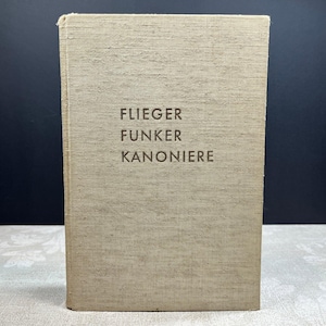 May include: A vintage book with a textured, tan cover. The title "FLIEGER FUNKER KANONIERE" is embossed in the center. The book is standing upright against a dark background, suggesting a historical or collectible item.