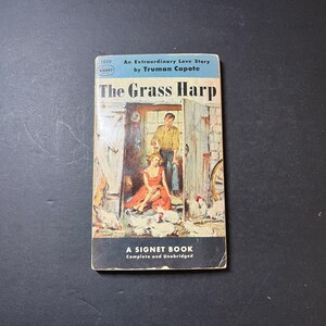 May include: A vintage Signet Book paperback edition of The Grass Harp by Truman Capote. The cover features a light blue top banner, a central illustration of a person in a red dress and a person in a yellow shirt in a barn, and a black bottom.