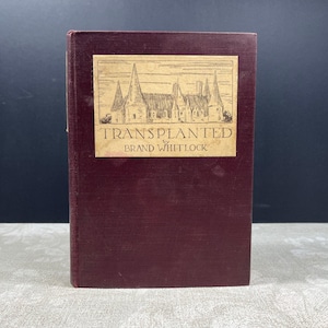 May include: A maroon hardcover book titled "Transplanted" by Brand Whitlock. The book's cover features a rectangular label with an illustration of a building with tall spires. The book is set against a dark background.