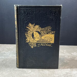 Op de afbeelding: Antiek hardcover boek getiteld "Darkness and Daylight in New York". De donkerblauwe kaft heeft gouden letters en illustraties. De rug is zichtbaar, met gouden details en slijtage.