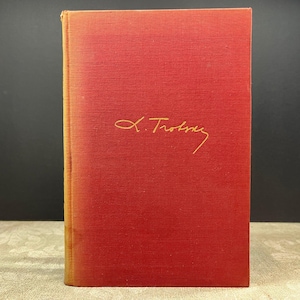 May include: A vintage book with a red cover and gold lettering. The title is "L. Trotsky" in a cursive font. The book has a textured surface and a slightly faded appearance, suggesting age.