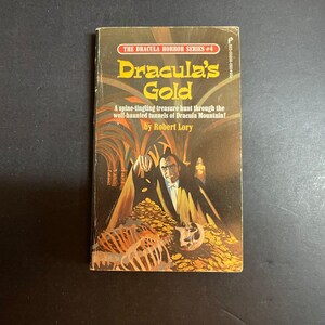May include: A paperback book titled Dracula's Gold by Robert Lory, part of The Dracula Horror Series #4. The cover features a dark illustration of a figure in a black cape standing over a pile of gold coins and a human skeleton in a tunnel.