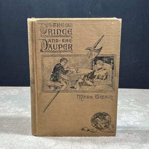 May include: A tan hardcover book titled The Prince and the Pauper by Mark Twain. The front cover features a black ink illustration of two figures seated at a table, a halberd, and a circular emblem. The text is printed in a decorative font.