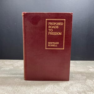 May include: A maroon hardcover book titled "Proposed Roads to Freedom" by Bertrand Russell. The title and author's name are in gold lettering within a gold rectangular frame on the front cover. The book is standing upright.