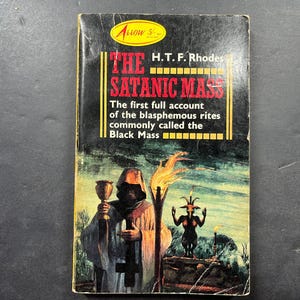 May include: A paperback book titled The Satanic Mass by H.T.F. Rhodes with a black cover featuring a hooded figure holding a chalice, a burning torch, and a horned figure standing on a stone wall against a dark, green-toned landscape background.