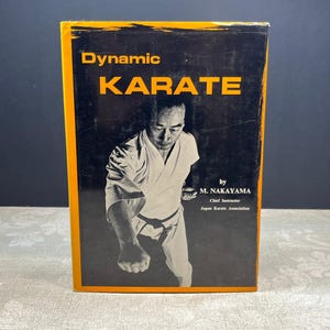 May include: A vintage book titled "Dynamic Karate" with a black and orange cover. The cover features a black and white photo of a man in a karate uniform performing a move. The author is M. Nakayama, Chief Instructor, Japan Karate Association.
