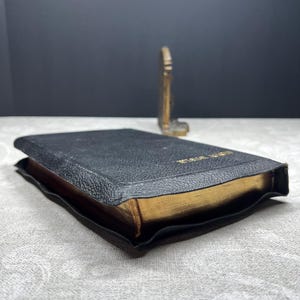 Puede incluir: Un libro encuadernado en cuero negro con bordes dorados. El libro est&aacute; abierto, revelando las p&aacute;ginas doradas. Un peque&ntilde;o objeto decorativo dorado est&aacute; en el fondo.