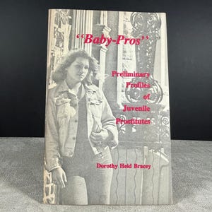 以下が含まれることがあります： ドロシー・ハイド・ブレイシー著『Baby-Pros: Preliminary Profiles of Juvenile Prostitutes』の白黒のブックカバー。カバーには、デニムジャケットを着た人物が装飾的な金属製の門の横に立っている写真が印刷されています。