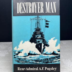 以下が含まれることがあります： リアー・アドミラルA.F.パグスリー著のヴィンテージ本「Destroyer Man」。表紙には、黒い背景と水色の背景に、太字の白い文字でタイトルが書かれた、海上の軍艦の白黒イラストが描かれています。