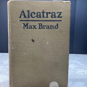 May include: A vintage book titled "Alcatraz" by Max Brand. The book has a faded olive green cover with black lettering. The cover shows signs of wear and tear, with some discoloration and minor damage.