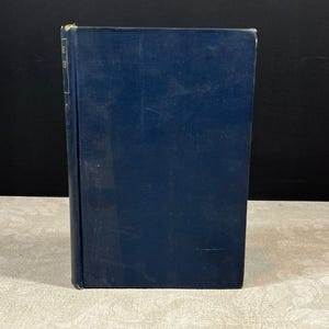 May include: A vintage hardcover book with a dark blue cover. The book is standing upright on a light-colored surface. The spine has a faded label. The book's cover shows signs of wear.
