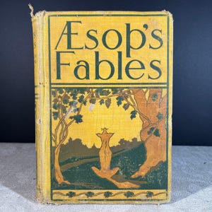 Op de afbeelding: Een vintage hardcover boek getiteld Aesop's Fables met een gele kaft, donkergroene tekst en een grafische illustratie van een vos die onder bomen zit. Het boek vertoont tekenen van slijtage langs de randen en de rug van het boek.