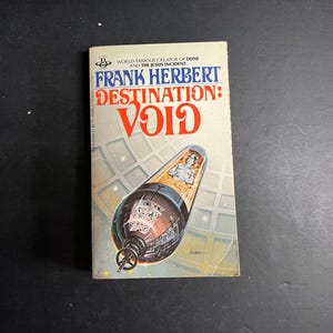 Puede incluir: Un libro de bolsillo titulado Destination: Void de Frank Herbert. La portada presenta un fondo crema con texto en azul y rojo, y una ilustración de una nave espacial cilíndrica con un interior metálico sobre un fondo espacial de cuadrícula.
