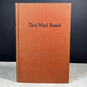 Könnte beinhalten: Ein Vintage-Buch mit einem strukturierten orangefarbenen Einband. Der Titel "Third Ward Newark" ist in schwarzer Schrift auf der Vorderseite aufgedruckt. Das Buch steht aufrecht vor einem dunklen Hintergrund.