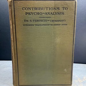 May include: An antique book titled "CONTRIBUTIONS TO PSYCHO-ANALYSIS" by Dr. S. Ferenczi, with "AUTHORIZED TRANSLATION BY DR. ERNEST JONES" printed below. The book has a faded olive green cover with black text and a black border.