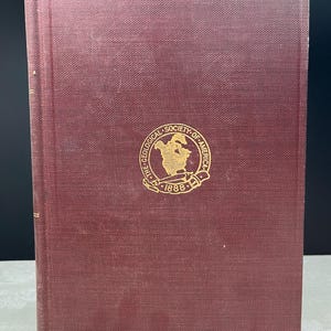May include: A maroon-colored book with a gold emblem of The Geological Society of America, established in 1888, is displayed. The book's cover has a textured appearance. The book is standing upright against a dark background.