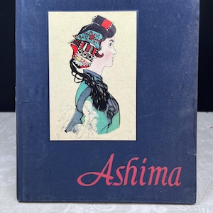 Puede incluir: Un libro azul marino con una ilustración enmarcada de una mujer de perfil con un tocado decorativo. La ilustración es en tonos verdes, rojos y negros. El título "Ashima" está escrito en rosa debajo.