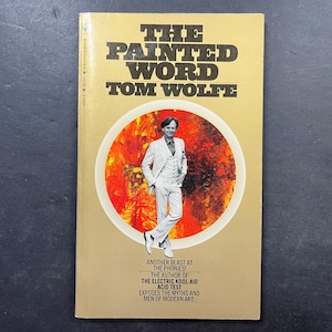 以下が含まれることがあります： トム・ウルフ著『The Painted Word』のペーパーバック。表紙はタンカラーで、中央には燃えるようなオレンジと赤の背景に白いスーツを着た人物の円形グラフィックが描かれています。テキストには「Another blast at the phonies!」と記載されています。