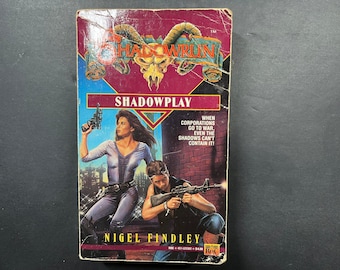 Può includere: Un libro tascabile intitolato Shadowrun: Shadowplay di Nigel Findley. La copertina presenta un logo con teschio cornuto, una persona in tuta blu con una pistola e un'altra in ginocchio con un fucile davanti a un muro di mattoni.