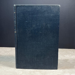 May include: A hardcover book with a dark navy blue cloth cover. The spine is visible on the left side, featuring faint, partially obscured gold lettering. The book is rectangular with sharp corners and a textured fabric surface on the front cover.
