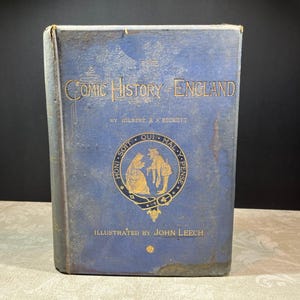 May include: A blue hardcover book titled Comic History of England by Gilbert A'Beckett, illustrated by John Leech. The front cover features gold embossed lettering and a central circular emblem depicting two figures with the text Honi Soit Qui Mal Y Pense.