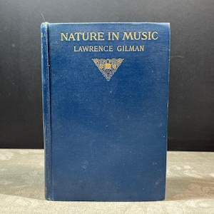 May include: A vintage book titled "Nature in Music" by Lawrence Gilman. The book has a dark blue cover with gold lettering and a decorative emblem. The book is standing upright against a dark background.
