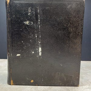 May include: A vintage black book with a textured cover. The book shows signs of wear, with scuffs and discoloration on the cover. The edges of the book are slightly worn, revealing the underlying material. The book has a rectangular shape and appears to be closed.